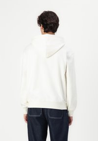 Sudadera color crema con capucha ajustable, textura suave y puños acanalados, combinada con jeans de mezclilla oscura, vista desde atrás.