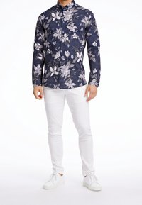 Camicia a fiori blu navy con fiori bianchi e grigi, maniche lunghe, abbinata a pantaloni bianchi a taglio slim e scarpe da ginnastica bianche.