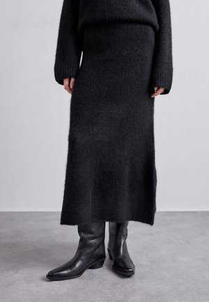 Personne portant une longue jupe noire duveteuse, un pull noir duveteux et des bottes en cuir noir mi-mollet, debout sur un sol gris.