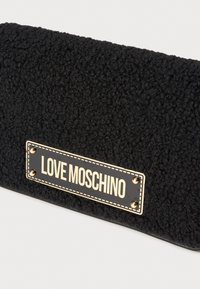 Clutch bag nera con texture, caratterizzata da una placca logo in oro con due borchie dorate. Esterno morbido e peloso, di forma rettangolare e profilo piatto.
