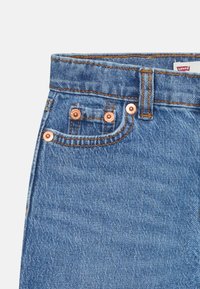 Jean en denim bleu clair avec des rivets en cuivre, fini avec des coutures orange contrastantes. Présente un design classique à cinq poches.