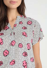Donna che indossa una camicetta bianca con pois neri e motivi floreali astratti rosa sparsi, che mostra la parte superiore del torso e parte del viso.