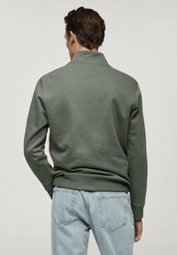 Felpa verde scuro con colletto, polsini e orlo a costine. Indossata con jeans azzurri, che mostrano un tessuto testurizzato e un design casual.