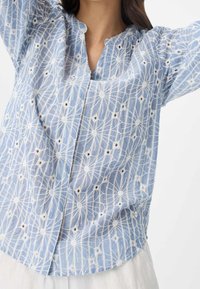 Blouse bleu clair avec broderies blanches présentant des motifs floraux et de petites touches brunes. Conception boutonnée avec un col arrondi.