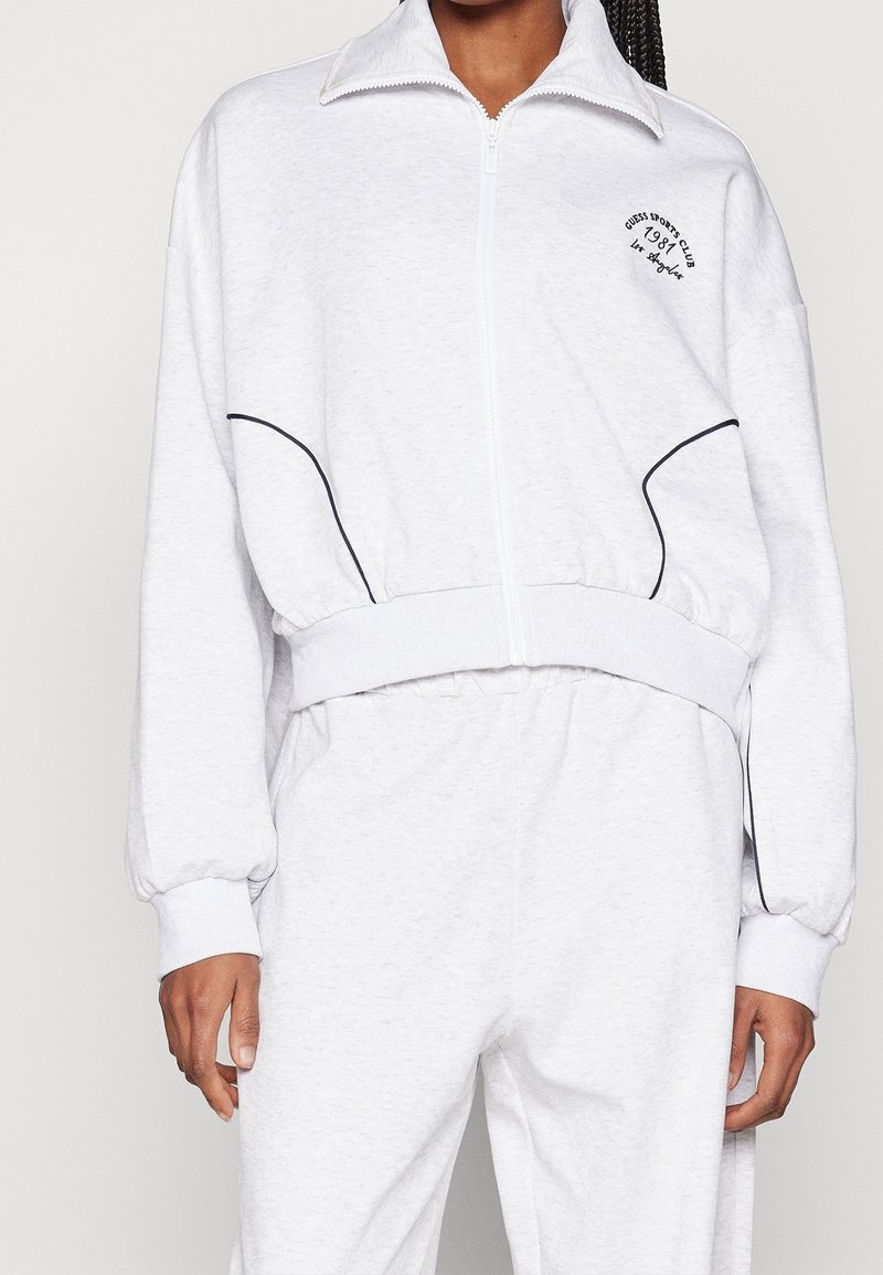 Sweat-shirt zippé gris clair en tissu doux avec col montant, poignets élastiques, accents en passepoil noir et détail logo sur la poitrine.