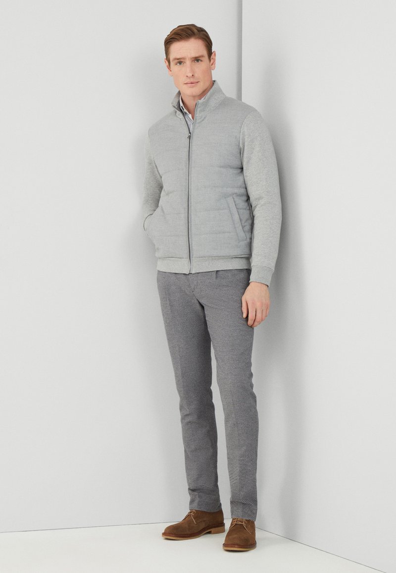 Mann steht an einer Eckewand und trägt eine hellgraue Zip-Jacke, karrierte Slim-Fit-Hosen und braune Wildlederschuhe.