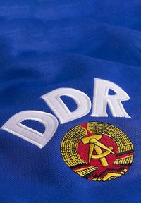 Blauwe stof met een opvallende witte geborduurde "DDR." Hieronder een rond embleem met een rode achtergrond, met een hamer, sikkel en tarwe.