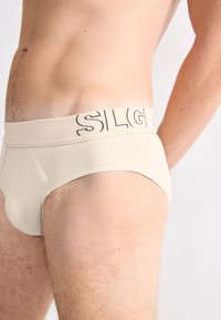 Beige Männerbriefs aus weichem Stoff mit glatter Textur. Mit breitem elastischem Bund, auf dem "SLIG" in Schwarz gedruckt ist.