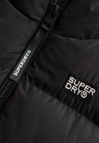 Czarna kurtka ocieplana o gładkiej fakturze, z zamkiem błyskawicznym z przodu oraz białą łatą z logo "SUPERDRY" na piersi.