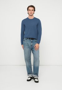 Niebieski, długi sweter z dzianiny z długim rękawem w połączeniu z jasnoniebieskimi, prostymi jeansami oraz czarnymi butami z białymi akcentami. Neutralne tło.