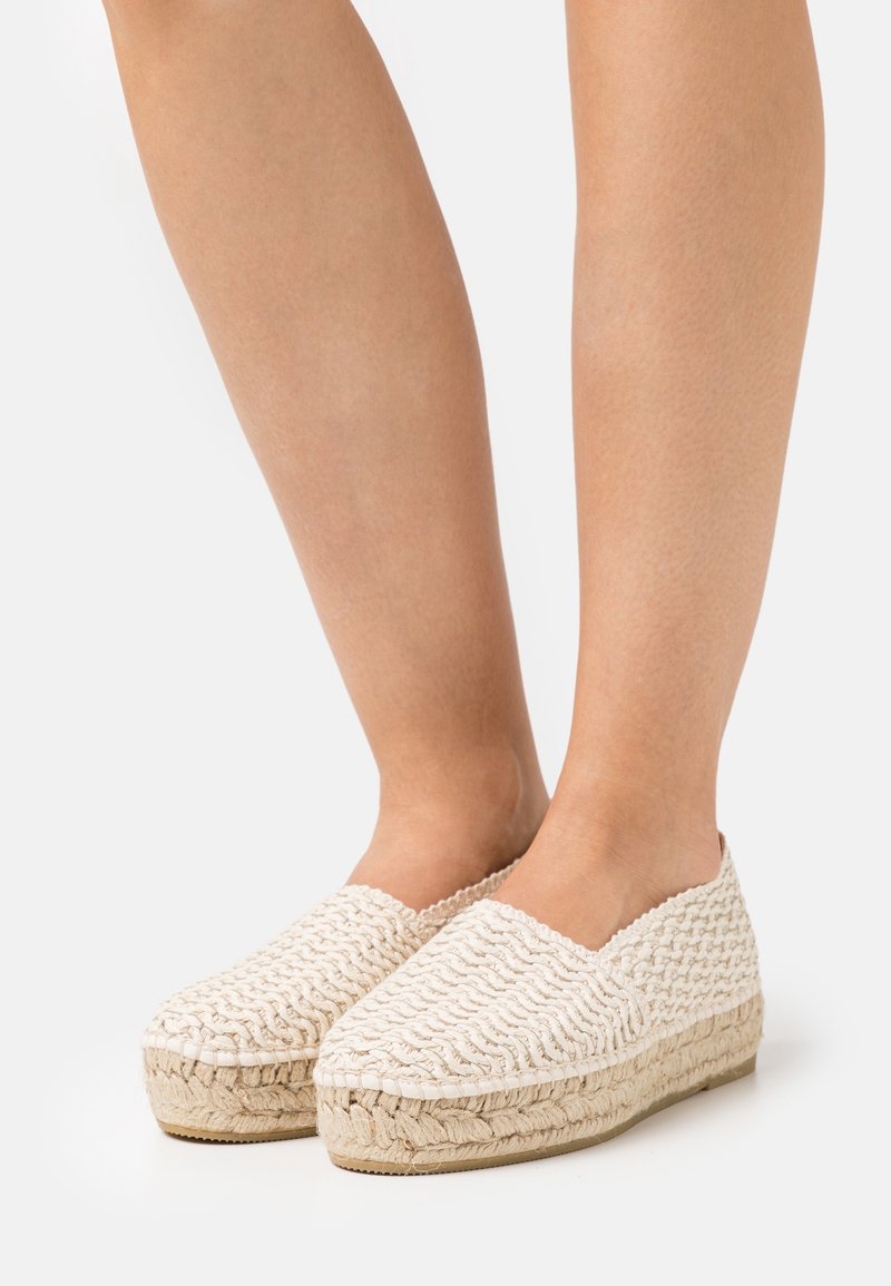 Pieds portant des espadrilles compensées tissées couleur crème avec semelle en jute tressé sur fond blanc.