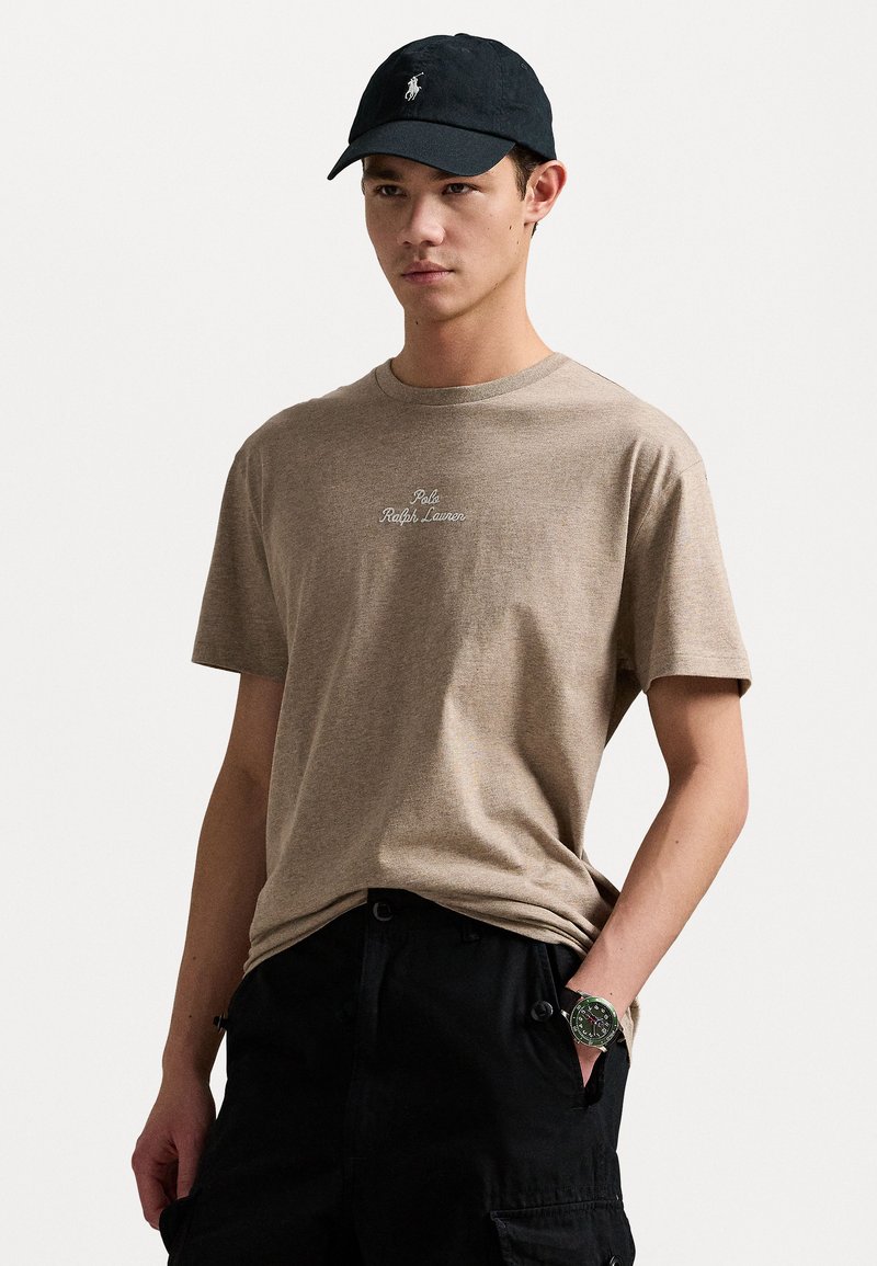 Καστανό βαμβακερό t-shirt με κεντητό λογότυπο "Polo Ralph Lauren", συνδυασμένο με μαύρο καπέλο και σκούρα cargo παντελόνια; διαθέτει κοντά μανίκια και στρογγυλή λαιμόκοψη.