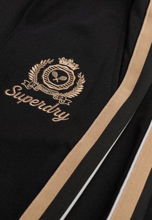 Tela negra con bordado dorado de "Superdry" y un diseño de laurel; acentuada con franjas beige y negras. Superficie texturizada.