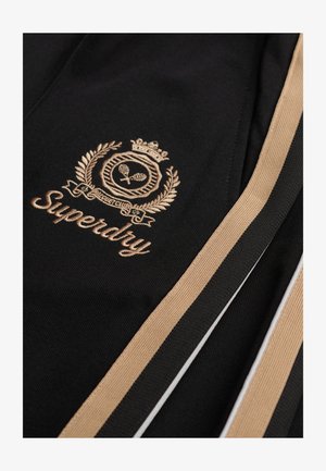 Tessuto nero con ricamo dorato della scritta "Superdry" e un design di alloro; accenti in strisce beige e nere. Superficie texture.