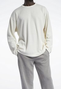 Osoba o ciemnej karnacji ubrana w fakturowaną, jasnobeżową koszulę z długim rękawem i szare spodnie, stoi z rękami w kieszeniach na jasnym tle.