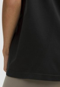 Sort atletisk top med en tekstureret overflade, der har en afrundet kant og subtile detaljeringer på sidepanelerne. Den er parret med lysfarvede leggings.