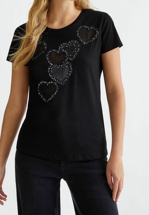 Femme portant un t-shirt noir à manches courtes avec six formes de cœur pailletées disposées en diagonale sur la poitrine, assorti à un jean foncé.