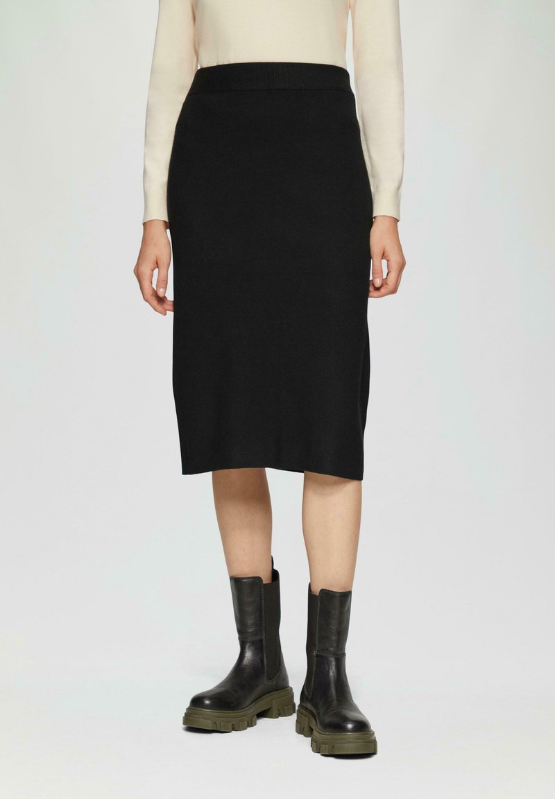 Zwarte geribde midi-rok met een aansluitende tailleband, gecombineerd met chunky zwarte leren enkellaarsjes met een olijfgroene zool.