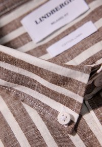 Camisa a rayas en marrón y crema con cuello, hecha de una mezcla de algodón y lino. Presenta un cierre con botones y etiquetas de la marca visibles por dentro.