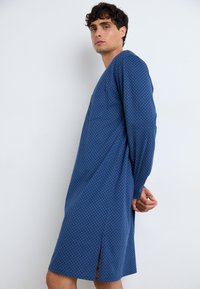 Blå, langermet kjole med geometrisk mønster, med V-hals og sideslits for bevegelsesfrihet, laget av et glatt stoff.