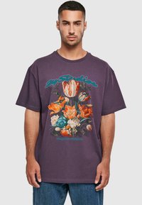 Lilla bomulls oversized t-skjorte med en fargerik blomstertrykk i oransje, blå og hvite blomster, som viser intrikate detaljer og tekstur.