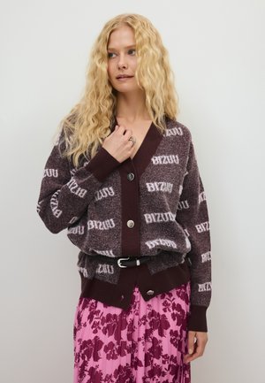 Vrouw met lang, krullend blond haar, een bruin patroonvest met knopen en een roze bloemen geplooide rok.
