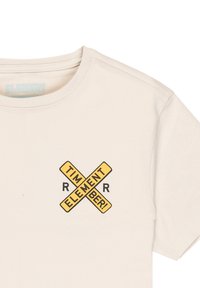Camiseta beige de cuello redondo con un gráfico de una señal cruzada amarilla que incluye las palabras "TIMBER! ELEMENT" y dos letras "R" en negro.
