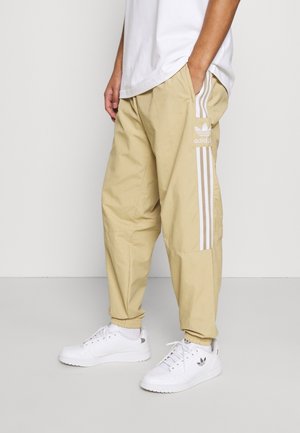 Persoon draagt beige Adidas trainingsbroek met witte strepen en logo, witte sneakers en een wit shirt, staand met handen in de zakken.