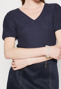 Tmavě modrý texturovaný top s výstřihem do V a nařasenými rukávy. Na světlé pleti je nápadný perlový náramek.