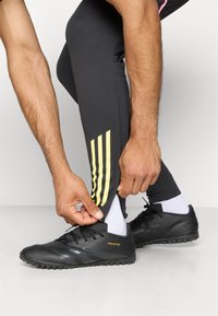 Svarte treningsko med tre striper, kombinert med tettsittende svarte leggings med gule striper nær ankelen. Nærbilde av hender som justerer buksene.