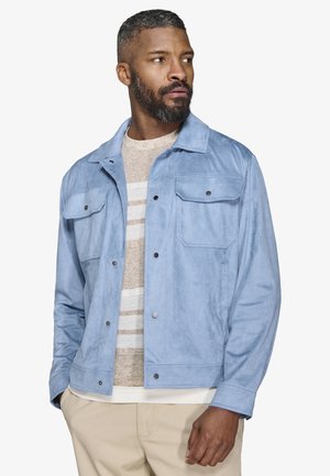 Mężczyzna o krótkich, kręconych włosach i brodzie, ubrany w jasnoniebieską zapinaną na guziki kurtkę, pod którą ma beżową, w paski koszulę oraz beżowe spodnie, patrzący w prawo.