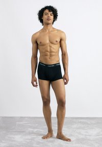 Boxeri negri din bumbac, cu un logo pe talie alb, design ajustat pe o suprafață texturată de culoare gri deschis, purtați de un model masculin.