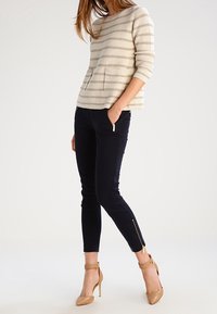 Maglione beige a righe con tasca frontale, abbinato a pantaloni slim-fit blu navy con cerniere laterali e scarpe con tacco color nude.