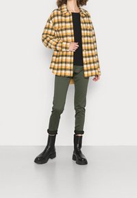 En gul och svart rutiga jacka över en svart topp, tillsammans med olivgröna byxor och svarta ankelboots. Jackan har en knappstängning fram.