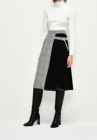 Woman wearing white ribbed turtleneck, black and plaid asymmetrical midi skirt with belt, and black knee-high heeled boots against white background.