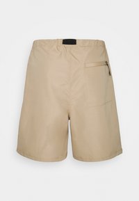 Short beige avec une taille élastique, une poche à fermeture éclair sur le côté droit, et une texture de tissu lisse et léger.
