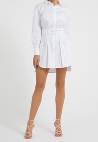 Abito camicia bianco a maniche lunghe con vita cintata, caratterizzato da pieghe e colletto appuntito, abbinato a tacchi strappy rosa chiaro.