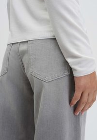 Graue Jeanshose mit tailliertem Bund, weitem Bein und zwei Gesäßtaschen, kombiniert mit einem hellen, langärmligen Hemd.
