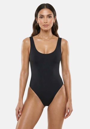 Femme aux longs cheveux foncés portant un maillot de bain noir une-pièce sans manches, debout face à un fond uni.