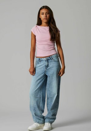 Jonge vrouw met lang donker haar, draagt een roze top met korte mouwen, losse lichtblauwe spijkerbroek en witte sneakers, staand tegen een grijze achtergrond.