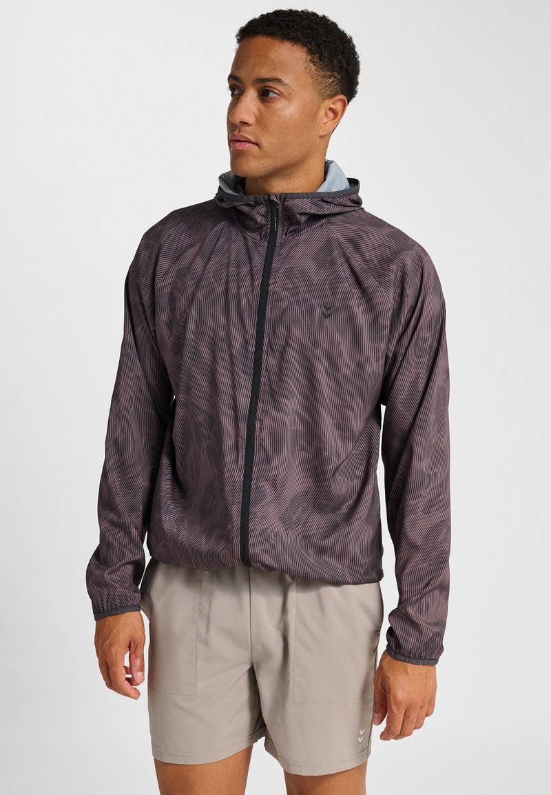 Leichte Herrenjacke in Dunkelviolett mit vertikalen Streifen, mit durchgehendem Reißverschluss, Kapuze und elastischen Bündchen. Kombiniert mit beigen Shorts.