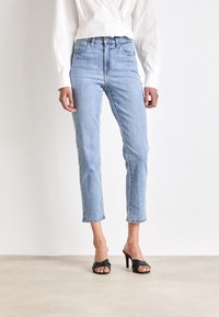 Kvinna i ljusblå högmidjade jeans som slutar ovanför anklarna, med svarta klacksandaler och en vit långärmad skjorta med knappar.