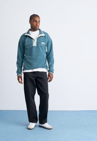 Een jonge man staat op een blauwe vloer, gekleed in een teal fleece trui, witte sneakers en zwarte broeken, kijkend naar links tegen een plain witte muur als achtergrond.