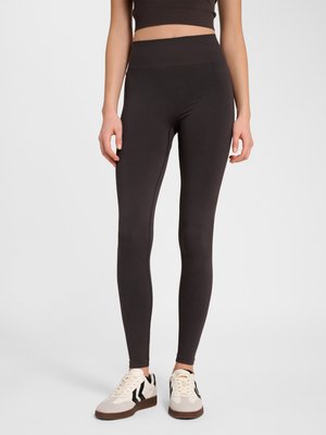 Vrouw draagt hoge taille zwarte legging en witte sneakers met zwarte strepen, staand tegen een effen lichte achtergrond.