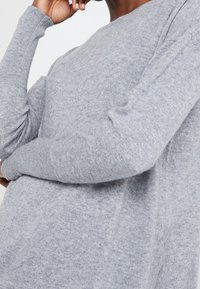 Pull en maille gris clair avec des manches longues, un col rond et un tissu texturé. Il présente des bords côtes aux poignets et à l'ourlet pour plus de détails.