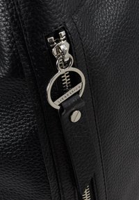 Gros plan d’une fermeture éclair d’un sac en cuir noir texturé avec une languette anneau argentée gravée du mot "LIEBESKIND."