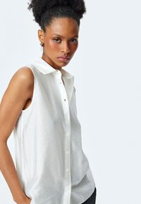 Ärmellose weiße Bluse mit durchgehender Knopfleiste und klassischem Kragen. Weicher, strukturiert wirkender Stoff mit leicht durchscheinendem Erscheinungsbild.