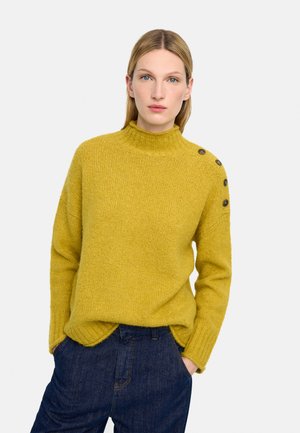 Kobieta w musztardowożółtym swetrze z dzianiny z trzema guzikami na ramieniu i ciemnoniebieskich dżinsach, stojąca z rękami w kieszeniach.