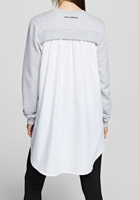 Persona che indossa una felpa grigia a maniche lunghe con un pannello di tessuto bianco sulla schiena e leggings neri, in piedi contro uno sfondo chiaro uniforme.