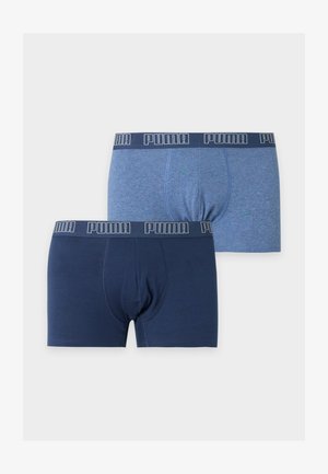Homme grand debout pieds nus, les bras croisés, portant un boxer Puma bleu foncé sur un fond clair uni.